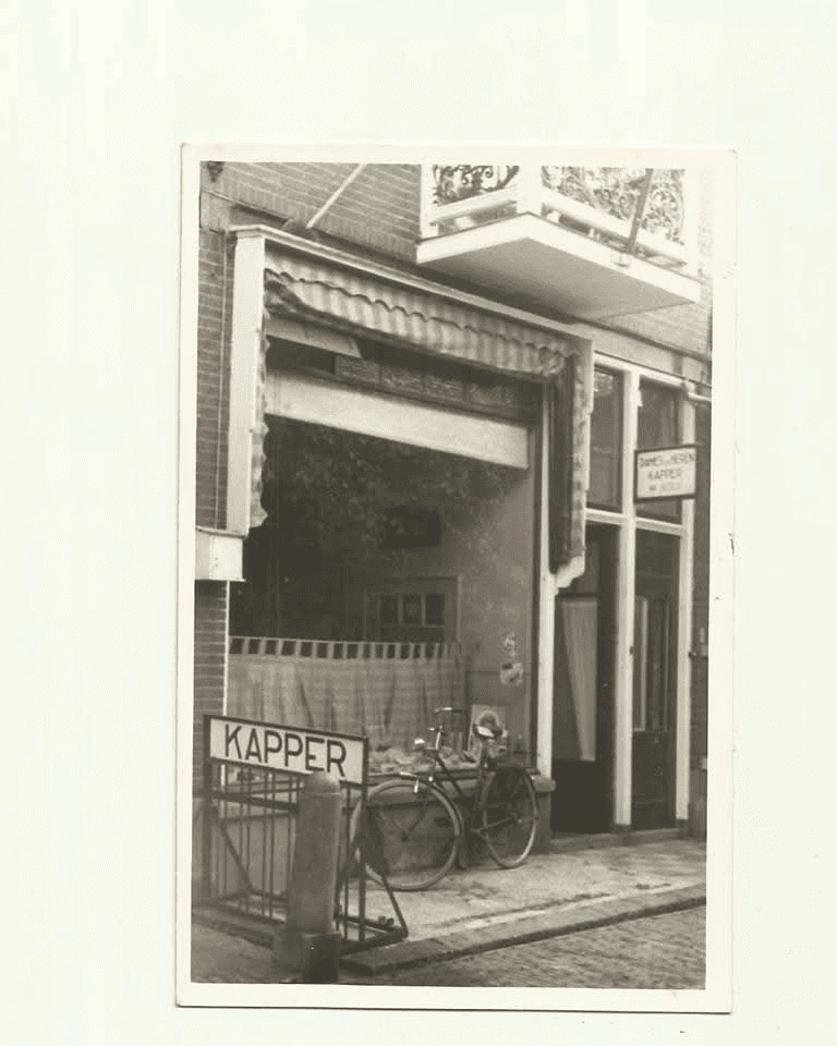 Historische foto van een kapperszaak, met een gestreept zonnescherm, een bord met Kapper en fietsen voor de deur.