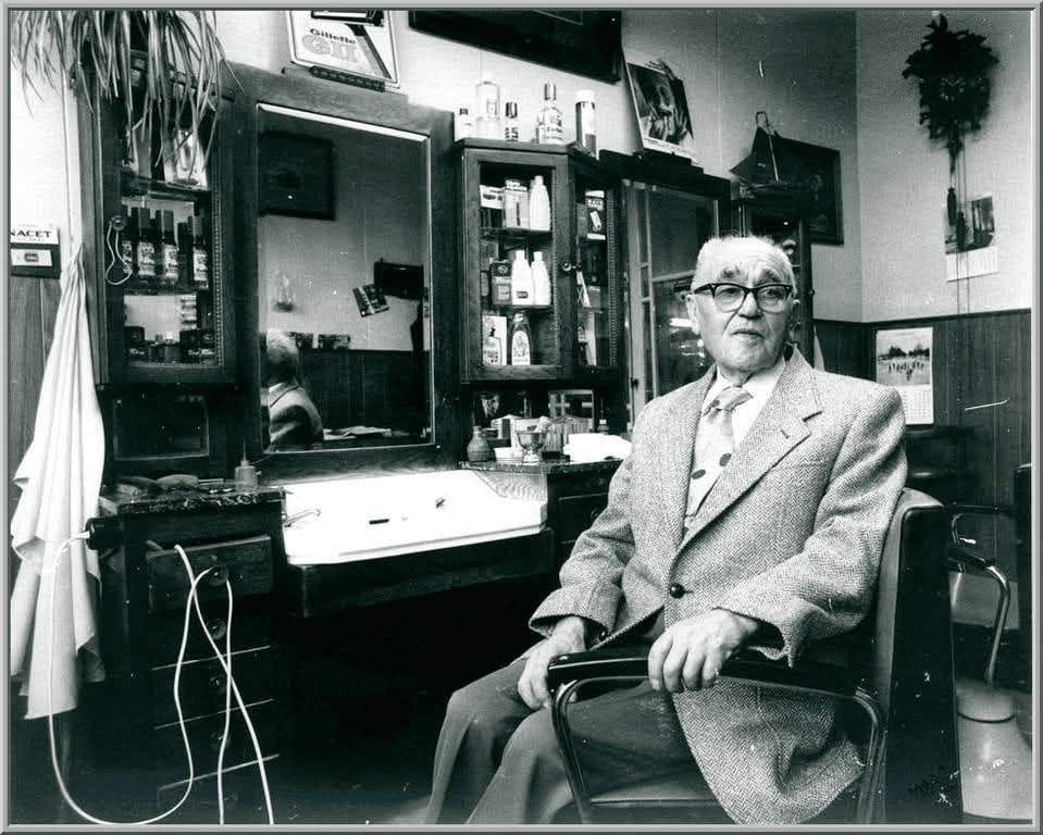 Een oudere man met bril zit in een kappersstoel in een salon met houten spiegels en vitrinekasten.